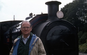 Image: 50-year-old white man with wire framed glasses wearing an open collared shirt and casual jacket, standing in front of a train engine.